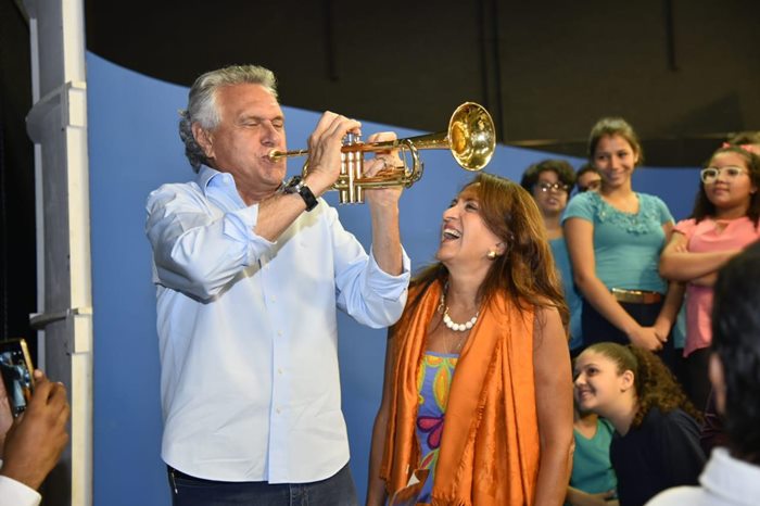 Governador Ronaldo Caiado ao lado da primeira-dama tocando trompete nos estúdios da ABC. Ao fundo estudantes da rede pública estadual de ensino.