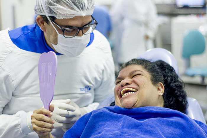 Paciente do Crer recebendo tratamento odontológico na unidade. Na imagem ele observa seu sorriso em um pequeno espelho, ao lado do dentista que a atendeu.