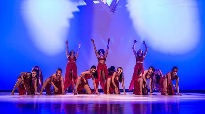 Doze bailarinas no palco interpretando o espetáculo: Empodere as Mulheres! em trajes vermelhos e sob luzes azuis.