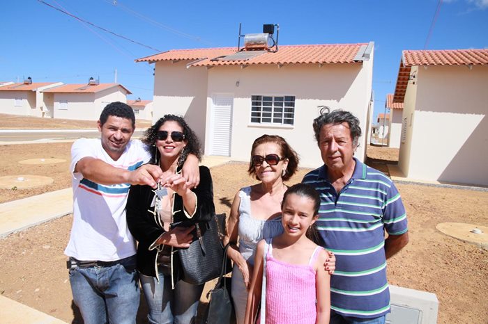 Família beneficiada com moradia no Residencial do Cerrado, em Anápolis. Na imagem um casal mais jovem,um casal da terceira idade e uma garotinha de aproximadamente  oio anos. Ao fundo as casas do residencial na cor bege.