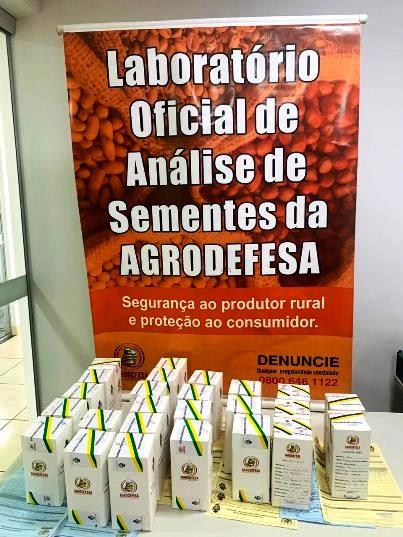 Banner do laboratório de sementes da Agrodefesa ao fundo e lotes lacrados de amostras de sementes a serem analisadas, em primeiro plano.