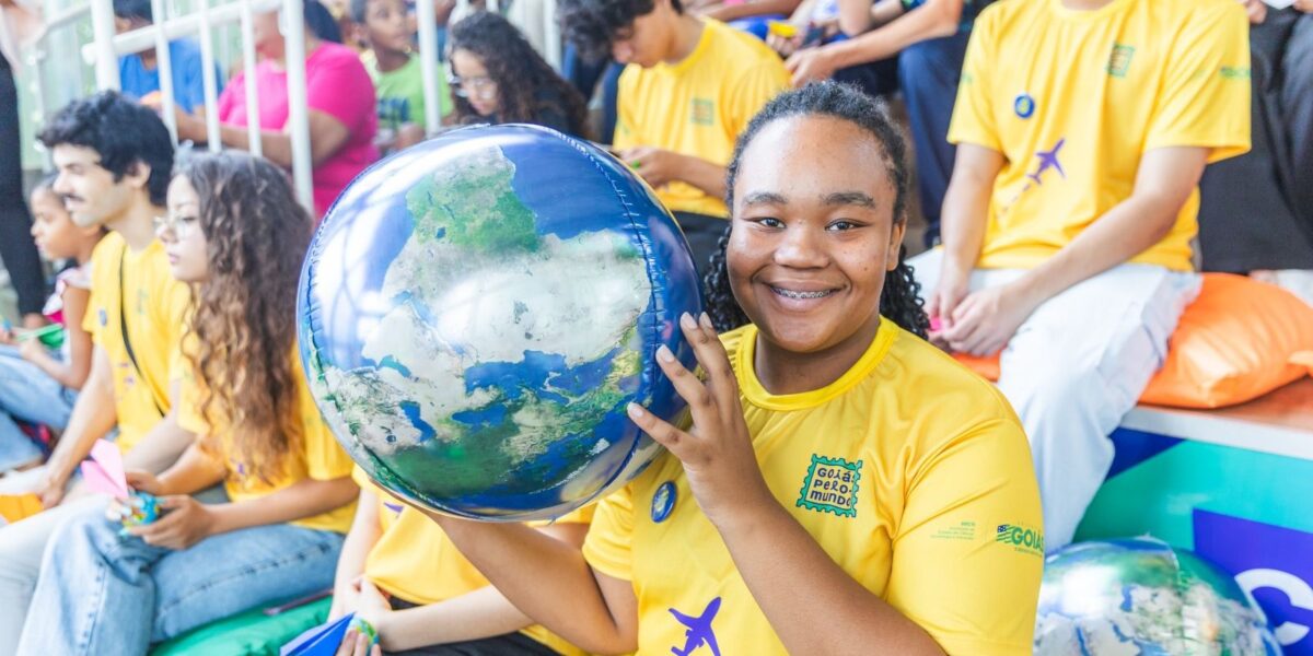 Goiás Social prorroga inscrições para intercâmbio de alunos da UEG e bolsistas ProBem - Garota negra de cabelo preso e camiseta amarela, segurando um globo nas mãos e sorrindo