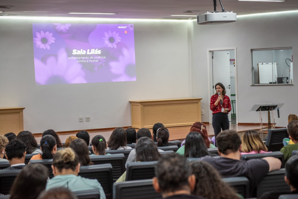 O Hospital Estadual de Urgências Governador Otávio Lage de Siqueira (Hugol) inaugurou, nesta quinta-feira (28/05), sua Sala Lilás, espaço destinado ao acolhimento e apoio de mulheres vítimas de violência. A iniciativa tem como objetivo fortalecer a assistência humanizada e garantir um atendimento mais seguro, reservado e acolhedor às vítimas atendidas. A Sala Lilás foi estruturada para oferecer privacidade, escuta qualificada e suporte durante o atendimento multiprofissional. O ambiente também poderá ser utilizado em atendimentos que que também envolvam crianças e adolescentes em situação de vulnerabilidade. O evento reuniu 168 profissionais do hospital, entre equipes assistenciais, multiprofissionais e setores estratégicos, reforçando a importância da qualificação contínua para o atendimento humanizado e identificação precoce de situações de violência. A inauguração contou com palestra voltada aos profissionais, ministrada pela coordenadora-geral dos Ciclos de Vida e Violências na Superintendência de Políticas e Atenção Integral à Saúde da Secretaria da Saúde de Goiás, Lígia da Fonseca Bernardes. Ela abordou temas relacionados ao enfrentamento da violência contra a mulher, incluindo: fluxo de atendimento dos casos; violência de gênero; Lei Maria da Penha; tipos de violência; ciclo da violência; identificação de sinais de abuso e vulnerabilidade. Capacitção das equipes Segundo Lígia, a capacitação das equipes de saúde é fundamental para garantir acolhimento adequado às vítimas. “Os profissionais da saúde têm papel essencial na identificação precoce dos sinais de violência e no acolhimento humanizado dessas mulheres”. “Muitas vezes, o hospital é a primeira porta de apoio procurada pela vítima, e estar preparado para esse atendimento pode fazer toda a diferença no rompimento do ciclo da violência”, destacou. Sala Lilás – avanço no cuidado De acordo com a supervisora multiprofissional do Hugol, Gabriela Vieira, a implantação da Sala Lilás representa um avanço importante no cuidado às vítimas em situação de vulnerabilidade. “A criação desse espaço fortalece o acolhimento humanizado dentro da unidade e proporciona mais segurança, privacidade e suporte emocional às pacientes atendidas. É um ambiente pensado para oferecer escuta qualificada e assistência integral”, afirmou. A criação da Sala Lilás integra as ações de fortalecimento das políticas de proteção e cuidado às vítimas de violência desenvolvidas pelo Hugol, promovendo um ambiente mais acolhedor e preparado para assistência integral aos pacientes em situação de vulnerabilidade. Além do acolhimento imediato, o espaço também busca contribuir para o encaminhamento adequado das vítimas à rede de apoio e proteção, fortalecendo o trabalho integrado entre saúde, assistência social e segurança pública. Saiba mais Sala Lilás do Hugol vai acolher mulheres vítimas de violência