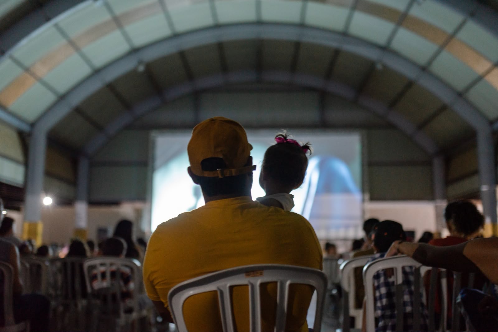 Governo de Goiás leva Cine Leitura do Bem a mais quatro municípios - público assistindo a sessão de cinema do projeto. Em primeiro plano, pai com bebê nos braços assistindo ao filme